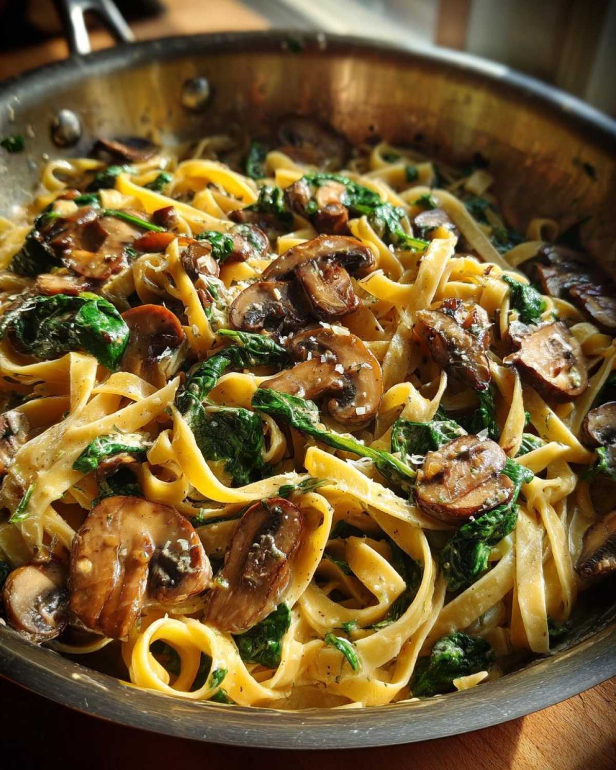 Close-up of creamy mushroom pasta featuring fettuccine, sautéed mushrooms, and wilted spinach in a stainless steel skillet.