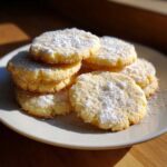 A stack of golden 3-ingredient butter cookies generously dusted with powdered sugar on a white plate.