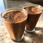 Two tall glasses filled with rich, blended frozen hot chocolate sitting on a sunlit countertop.