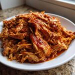 A close-up of saucy, shredded pulled chicken piled high on a white plate.