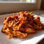 A close-up of saucy, shredded pulled chicken piled high on a white square plate.