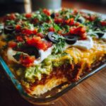 A close-up view of a serving dish containing the amazing 7 layer dip, showing layers of ingredients topped with guacamole, sour cream, tomatoes, olives, and cilantro.