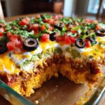 Close-up of a delicious 7 layer dip in a glass baking dish, showing layers of meat, cheese, guacamole, sour cream, topped with tomatoes, olives, and cilantro.
