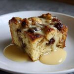 A close-up of a moist slice of bread pudding, studded with raisins and drizzled generously with vanilla custard sauce.