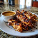 Close-up of perfectly grilled chicken satay skewers served on a white plate next to a small bowl of creamy peanut sauce.