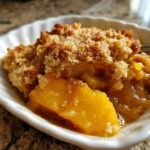 Close-up of a warm serving of peach cobbler in a white dish, showing juicy fruit and a golden crumb topping.
