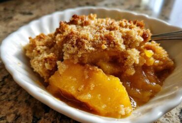 Close-up of a warm serving of peach cobbler in a white dish, showing juicy fruit and a golden crumb topping.