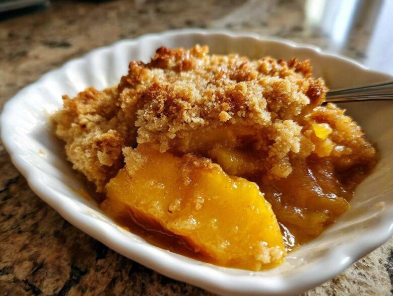 Close-up of a warm serving of peach cobbler in a white dish, showing juicy fruit and a golden crumb topping.