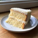A close-up of a moist slice of vanilla cake recipe layered with white frosting, served on a small white plate.