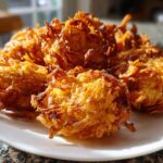 A close-up of several golden brown, crispy Amish Onion Fritters piled on a white plate, catching bright sunlight.