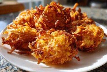 A close-up of several golden brown, crispy Amish Onion Fritters piled on a white plate, catching bright sunlight.