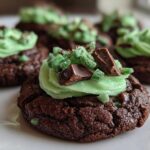 A close-up of a rich, dark chocolate Andes Mint Cookies topped with bright green frosting and chopped chocolate pieces.