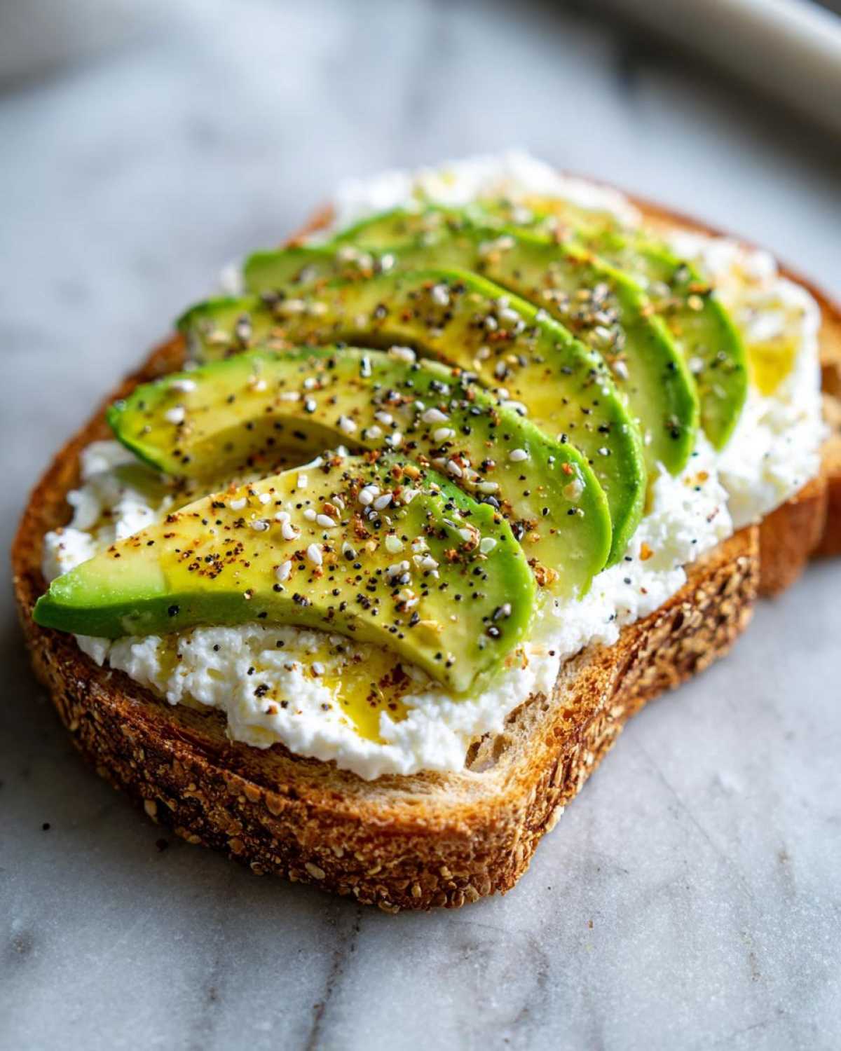 Close-up of a slice of whole grain cottage cheese toast topped with fanned avocado slices and everything bagel seasoning.