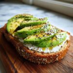 A slice of toasted bread topped with cottage cheese toast, layered with sliced avocado and everything bagel seasoning.