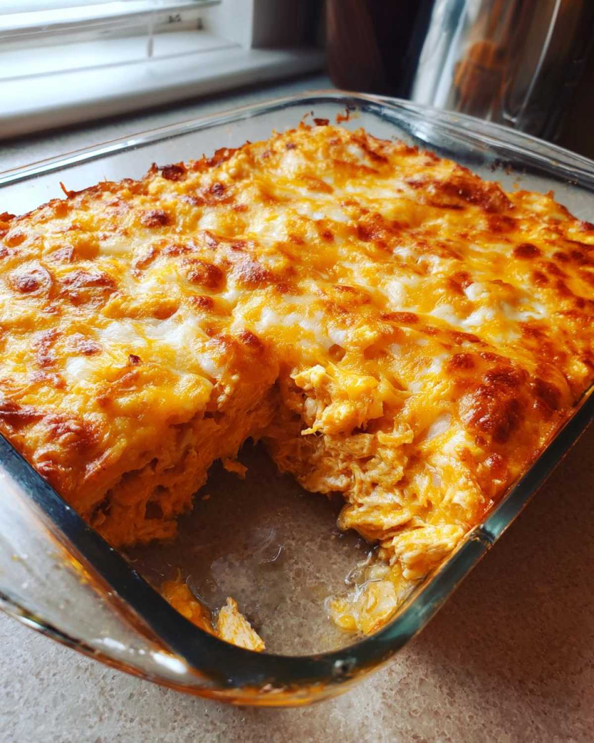 A square glass baking dish filled with hot chicken enchilada dip, showing a scoop removed to reveal shredded chicken texture.