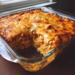 A close-up of baked chicken enchilada dip in a glass dish, showing shredded chicken mixture and a bubbly, golden-brown cheese crust.