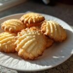 A close-up of several golden brown butter cookies with ridged tops stacked on a white plate, illuminated by sunlight.