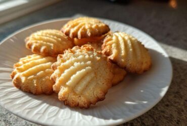 A close-up of several golden brown butter cookies with ridged tops stacked on a white plate, illuminated by sunlight.