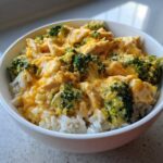 A close-up of a white bowl filled with fluffy white rice topped with creamy, cheesy chicken and bright green broccoli florets, representing cheesy chicken and rice.