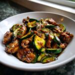 Close-up of a white bowl filled with glossy chicken zucchini stir fry, coated in a dark sauce.