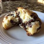 A chocolate chip cheesecake cookie broken in half showing the creamy cheesecake filling and melted chocolate chips.