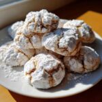 A pile of freshly baked cool whip cookies heavily coated in white powdered sugar, sitting on a white plate.