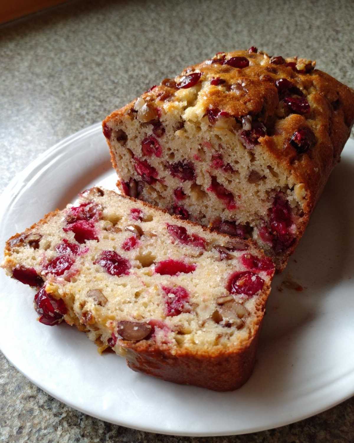 A close-up of a loaf of cranberry walnut bread, with one slice cut and resting against the main loaf, showing juicy cranberries and walnuts.