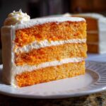 Close-up of a three-layer slice of bright orange Creamsicle Cake filled and frosted with white cream cheese icing.