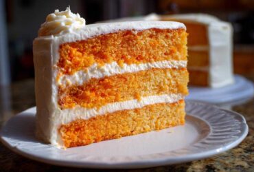 Close-up of a three-layer slice of bright orange Creamsicle Cake filled and frosted with white cream cheese icing.