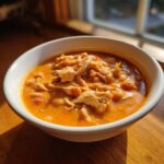 A close-up of a white bowl filled with creamy, orange-hued buffalo chicken chili topped with shredded chicken and beans.