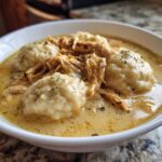 Close-up of a white bowl filled with creamy chicken and dumpling soup, featuring shredded chicken and fluffy dumplings.