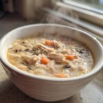 A steaming white bowl filled with creamy chicken rice soup featuring shredded chicken, carrots, and wild rice.