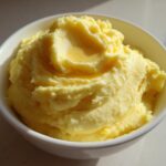 A close-up of perfectly creamy mashed potatoes, swirled high in a white bowl with melted butter pooling on top.