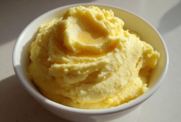 A close-up of perfectly creamy mashed potatoes, swirled high in a white bowl with melted butter pooling on top.
