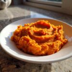 A close-up of vibrant orange, creamy mashed sweet potatoes served in a white bowl.