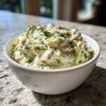A white bowl filled with chunky, creamy pickle dip, garnished generously with fresh dill.