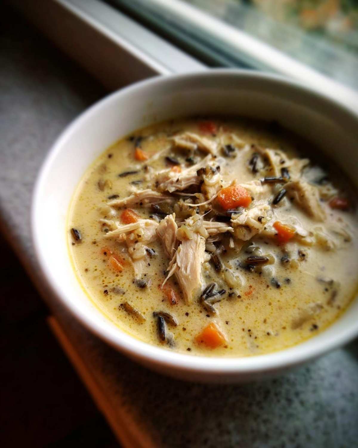 A close-up of creamy rice soup featuring shredded chicken, wild rice, and diced carrots in a white bowl.