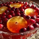 Close-up of a glass punch bowl filled with red Thanksgiving punch, garnished with floating orange slices and fresh cranberries.
