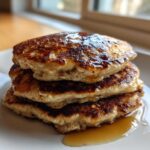 A close-up of a stack of three fluffy oatmeal pancakes drizzled generously with maple syrup.
