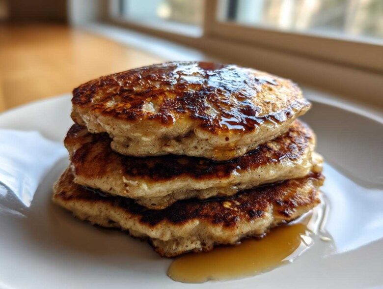 A close-up of a stack of three fluffy oatmeal pancakes drizzled generously with maple syrup.