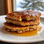 A close-up stack of three fluffy oatmeal pancakes generously topped with syrup on a white plate.