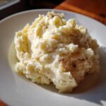 A serving of fluffy roasted garlic mashed potatoes with visible specks of roasted garlic on a white plate.