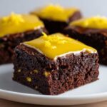 A close-up of a fudgy chocolate brownie square topped with bright yellow lemon glaze and zest, part of a batch of lemon brownies.