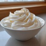 A white bowl filled with perfectly piped, glossy white sugar cookie icing, set near a window.