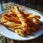 A pile of freshly baked, flaky, golden brown cheese straws dusted with seasoning, served on a white plate.