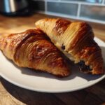 Two freshly baked, glossy chocolate croissant pastries resting on a white plate on a wooden countertop.