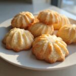 A close-up of several golden, piped butter cookies arranged on a white plate, catching bright sunlight.