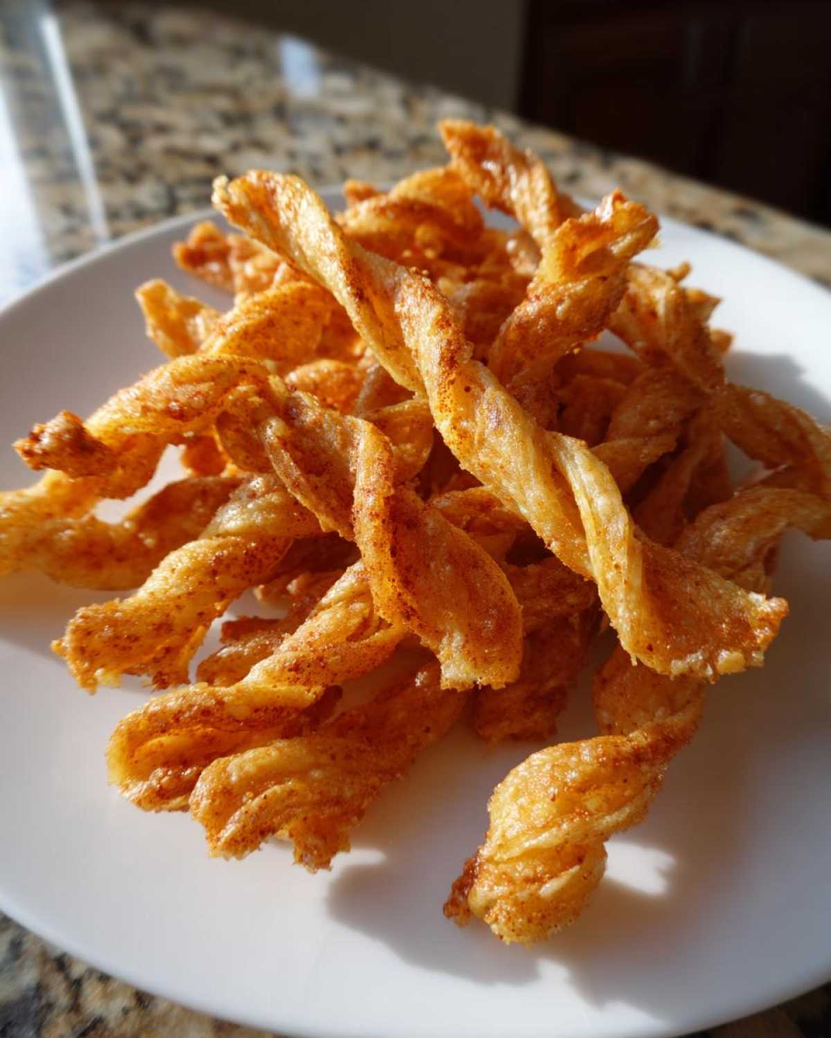 A pile of golden, flaky, twisted cheese straws seasoned with paprika or chili powder, resting on a white plate.