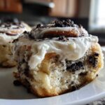 Close-up of a gooey Oreo cinnamon roll covered in white frosting and crushed cookie pieces.