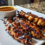 Close-up of perfectly grilled chicken satay skewers served on a white plate next to a small ramekin of creamy peanut sauce.